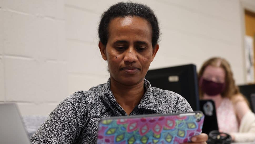 Adult student focused on a colorful tablet during a class session, with another student working at a computer in the background.