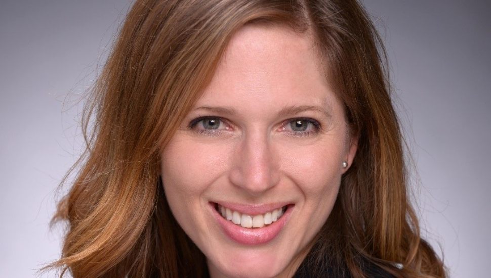 Headshot of Anna Brown: Light brown hair, smiling, against a grey-colored backdrop.