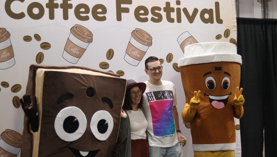 Two attendees pose with chocolate and coffee cup mascots in front of a 'Coffee Festival' backdrop decorated with coffee beans and cups at the Pennsylvania Chocolate and Coffee Festival.