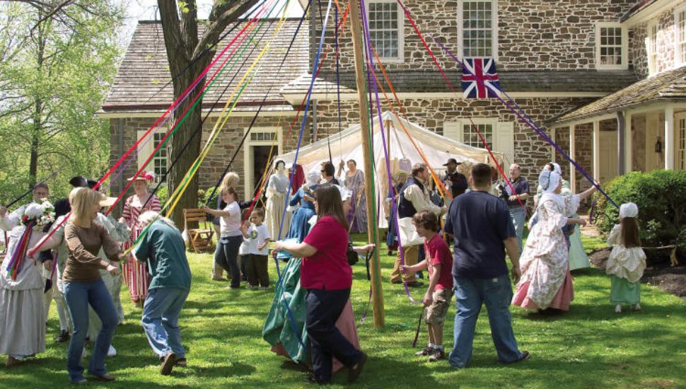 Families gather around the Maypole for dancing. It is a sunny day and there is bright green grass. Families and kids hold onto brightly colored ribbons around the maypole. There are vendors and crafters at tables.