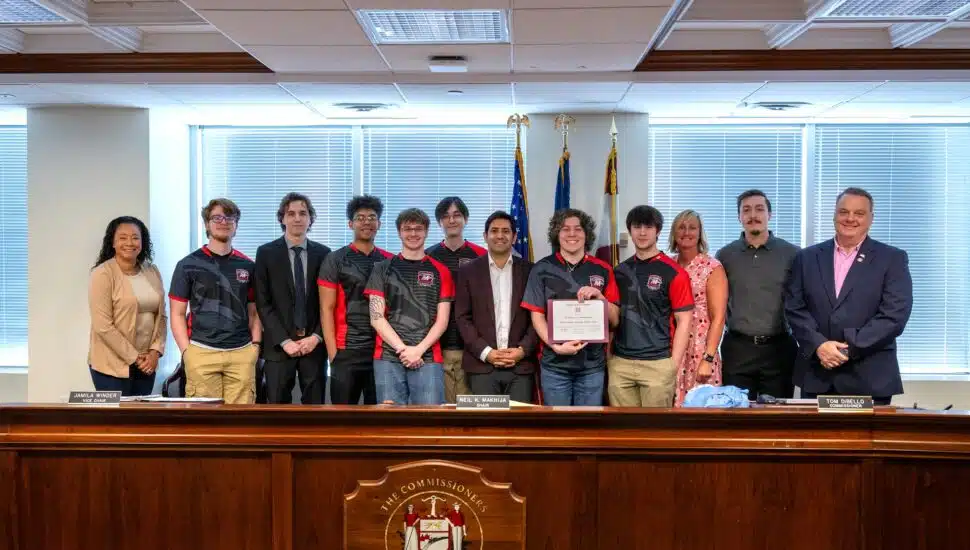 Members of the Montgomery County Community College Mustangs eSports team pose with Montgomery County Commissioners and staff after receiving a certificate of recognition for winning the NJCAAE national championship, standing behind a wooden dais with official seals and flags in the background.