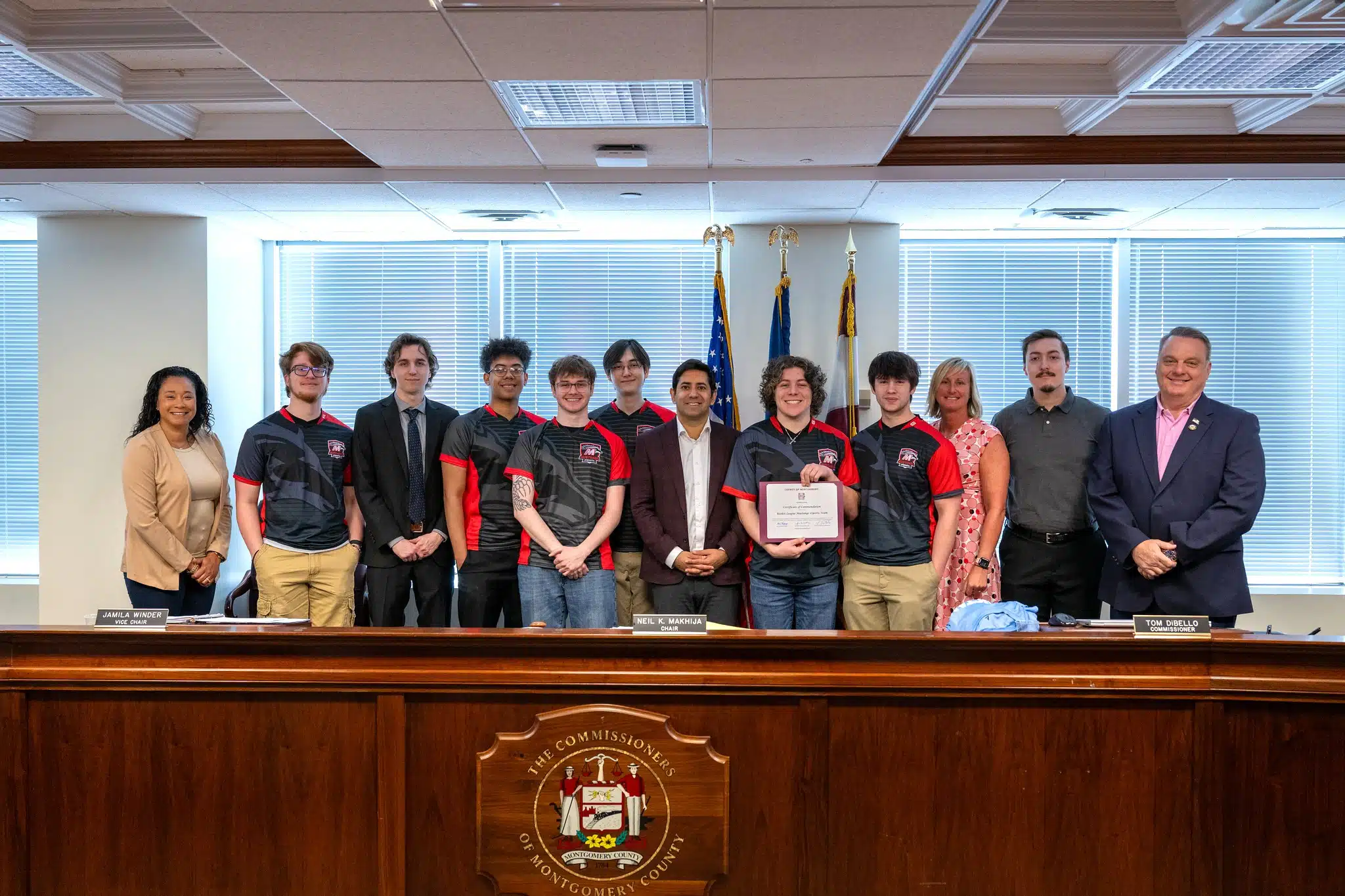 Members of the Montgomery County Community College Mustangs eSports team pose with Montgomery County Commissioners and staff after receiving a certificate of recognition for winning the NJCAAE national championship, standing behind a wooden dais with official seals and flags in the background.