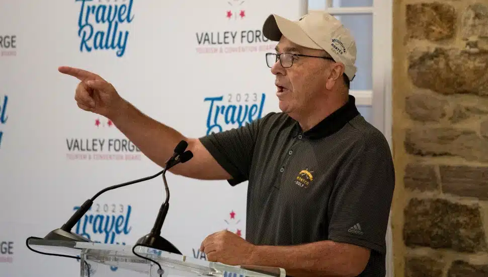 Mike Bowman, wearing a Montco Golf polo and cap, speaks passionately at a podium during the 2023 Travel Rally hosted by the Valley Forge Tourism & Convention Board.