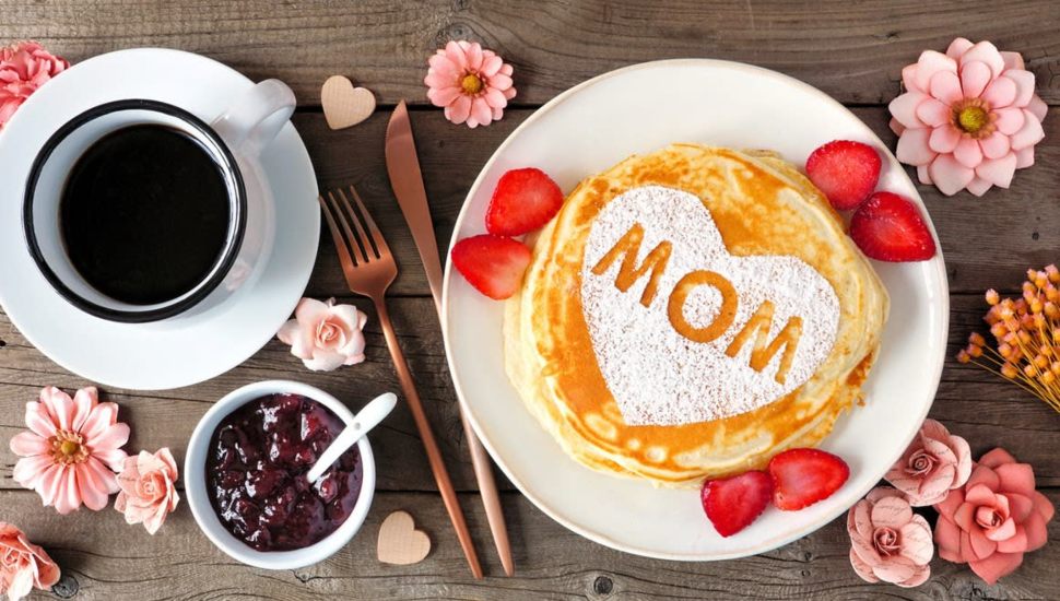 A photo of breakfast items/meal and flowers with the word "mom" written on a plate of pancakes.