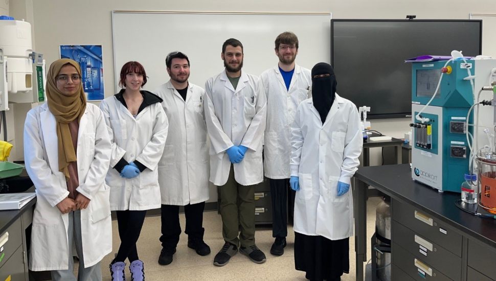 Group photo of six Montgomery County Community College Biotechnology students wearing white lab coats, standing together in a classroom setting.