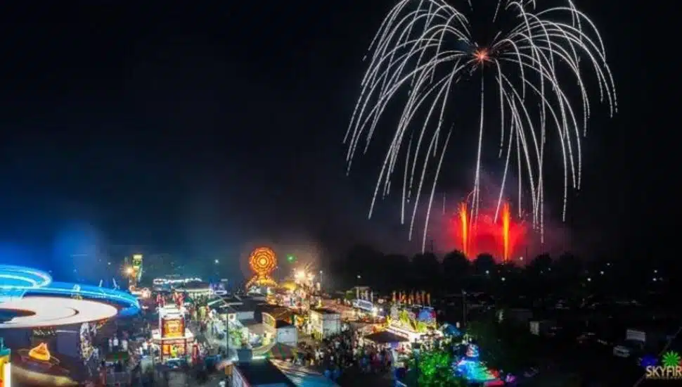 Nighttime carnival scene with bright rides, game booths, and a large crowd, as vibrant fireworks light up the sky during a 4th of July celebration in Montgomery County, PA.