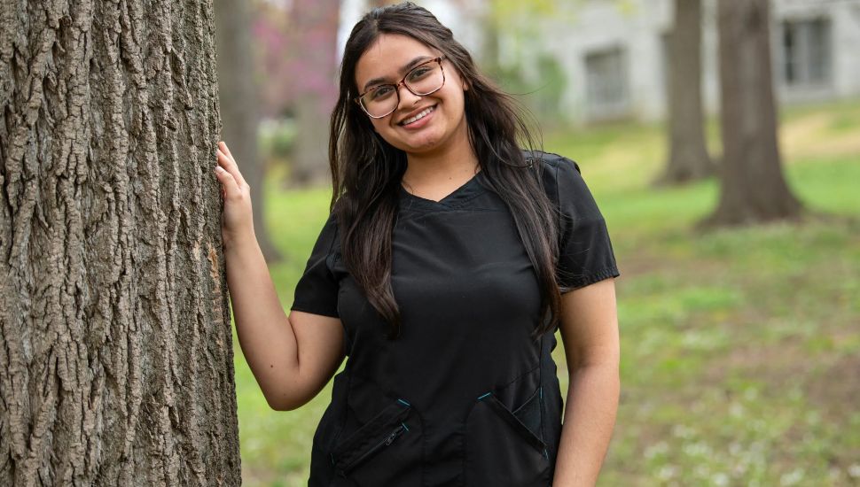 Afreen Snaha pictured: She's a young woman in black medical scrubs smiling and standing next to a tree in a park-like setting with buildings in the background.