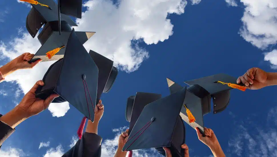 The students holding a shot of graduation cap by their hand in a bright sky.