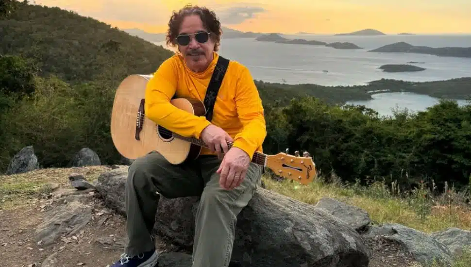 John Oates sits with his guitar on a hill overlooking the water.