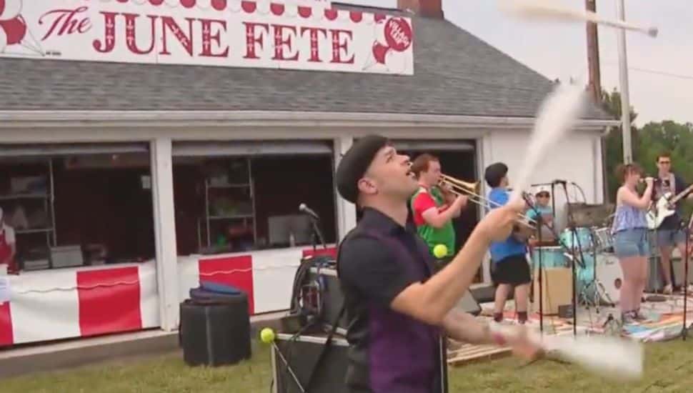 A man juggles bowling pins in front of the June Fete sign.