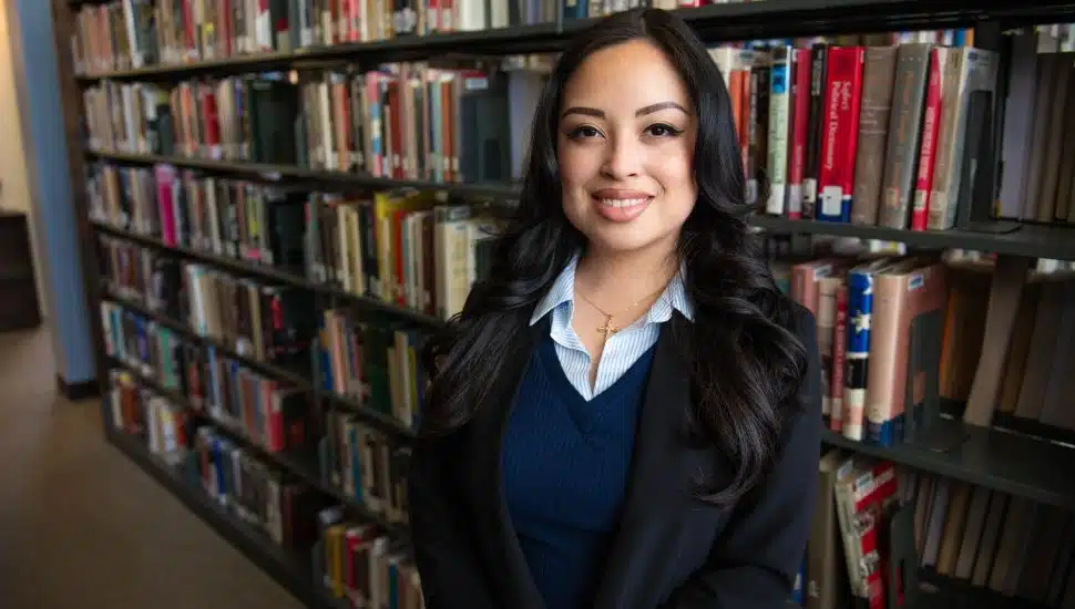 Headshot of Kimberly Alfaro Jimenez in a library setting, with a background of rows of books. She has long dark hair and wears navy and black, smiling.