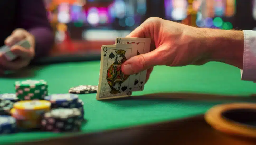 A hand holding pocket Queen and King at a poker table.