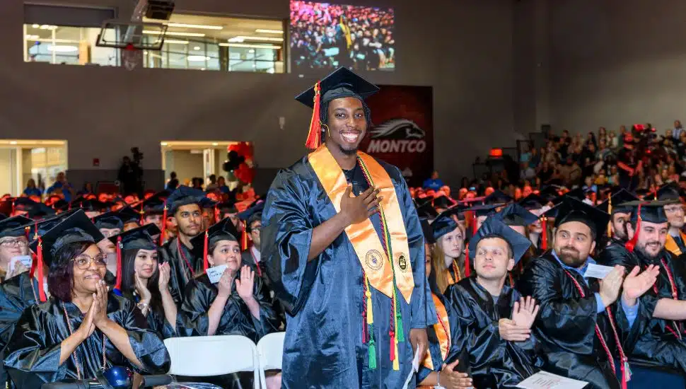 Josh Reid in a cap and gown stands smiling and proudly holding his hand to his chest during a commencement ceremony. He wears multiple honor cords and an orange stole. Other graduates in black gowns are seated around him, clapping and smiling. The ceremony is held indoors, with a large audience in the background. A screen behind displays part of the event and the word "MONTCO" with a mustang logo.
