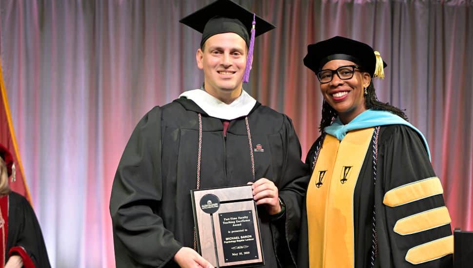 Graduating student Michael Baron, in cap and gown, holds a plaque while standing next to a smiling Dr. Chae Sweet in academic regalia at a graduation ceremony.