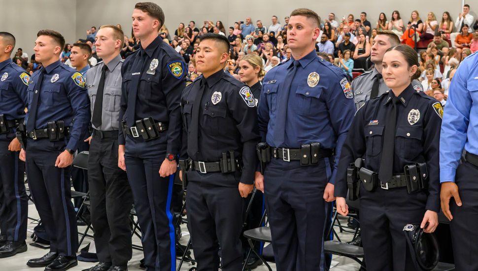 Police academy cadets in uniform stand in formation during a graduation ceremony, with a seated audience watching in the background.