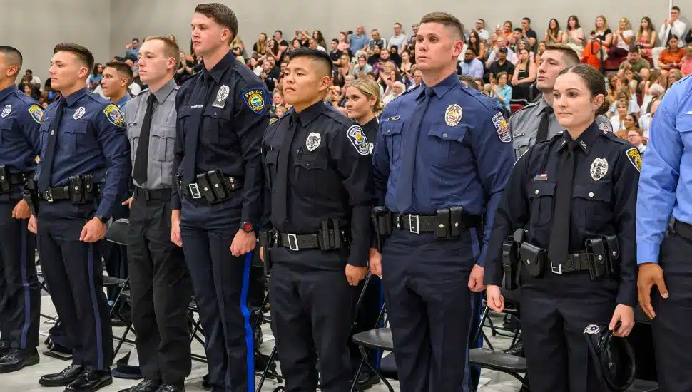 Police academy cadets in uniform stand in formation during a graduation ceremony, with a seated audience watching in the background.