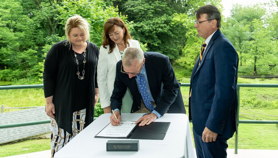 West Chester University and East Whiteland Township held a formal signing ceremony to celebrate their historic partnership in preserving a 52-acre farm that was gifted by an anonymous donor. Pictured (L to R) are West Chester University President Laurie Bernotsky, Executive Director of the West Chester University Foundation Deb Cornelius, East Whiteland Township Manager Steve Brown (signing), and Chairman of the East Whiteland Township Board of Supervisors Scott Lambert. A man in a suit signs a document on a table outdoors, while three other professionally dressed individuals—two women and one man—stand beside him, observing. The scene is set against a backdrop of lush green trees, suggesting a formal signing event in a natural setting.