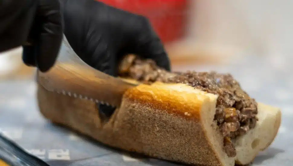 a hand in a black glove cuts through a cheesesteak.