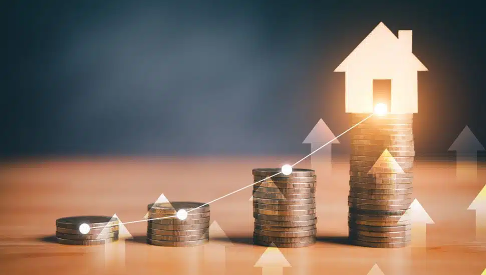 Stacks of coins with glowing house icon and upward arrows, symbolizing real estate investment growth.