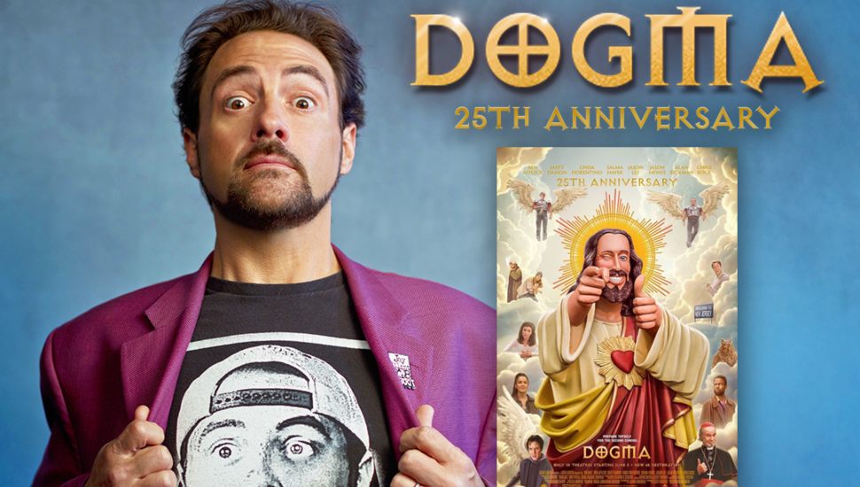 Filmmaker Kevin Smith poses pulling open his blazer to reveal a Silent Bob shirt, standing next to a poster promoting the 25th anniversary of his film Dogma, featuring a stylized image of Buddy Christ surrounded by characters from the movie.