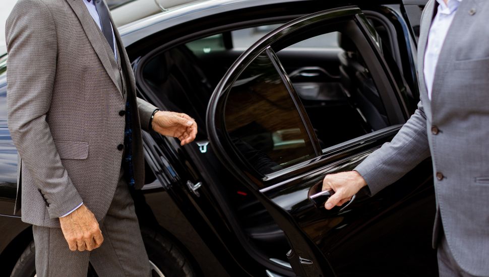 A businessman is greeted by his chauffeur as he approaches a black car.