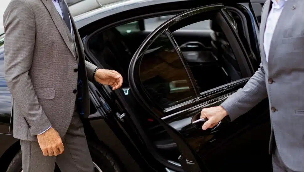 A businessman is greeted by his chauffeur as he approaches a black car.