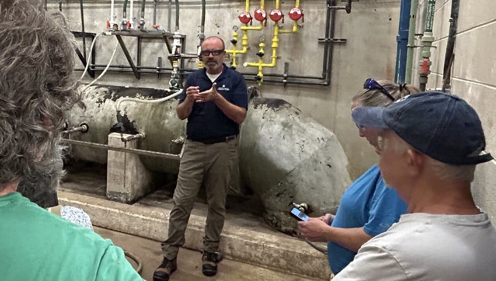 A man in a dark polo shirt and khaki pants gives a tour to a small group inside an industrial facility, standing in front of large cylindrical tanks and an array of pipes and valves. Several people listen attentively, one checking a phone. The setting appears to be a water treatment plant or similar utility facility.