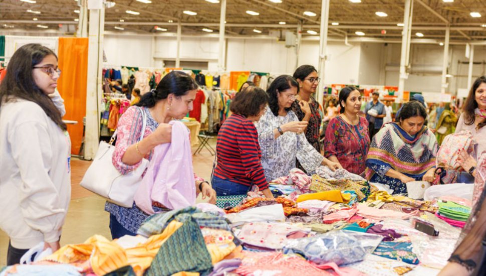 Visitors browse colorful traditional South Asian clothing and fabrics at a vendor booth during the Heritage India Festival inside a large expo hall.
