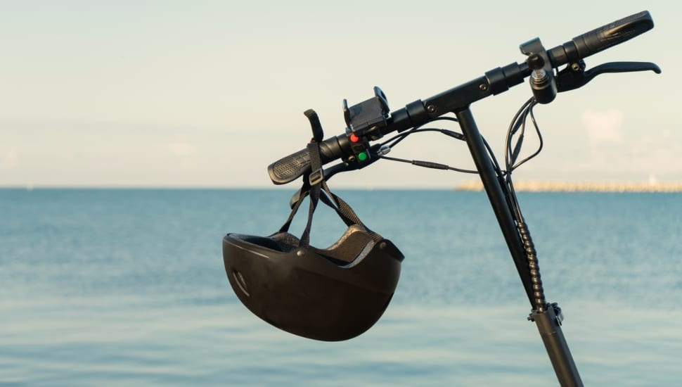 A safety helmet hanging on the handlebar of a an e-scooter near the ocean.