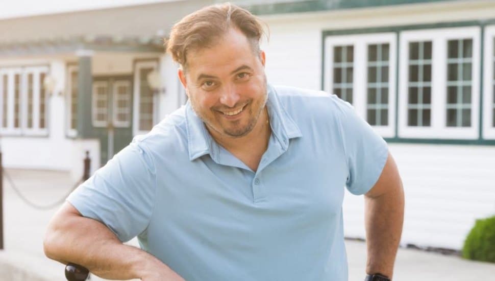 Man smiling outdoors in a light blue polo shirt, leaning casually on a railing in front of a white building with green trim and large windows.