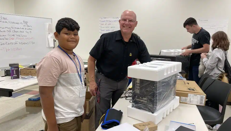 A middle school student and an instructor smile next to a partially unpacked computer tower during Montgomery County Community College’s PC Creation Station summer camp. In the background, other students work on assembling their own PCs in a classroom setting with boxes and computer parts spread across tables.