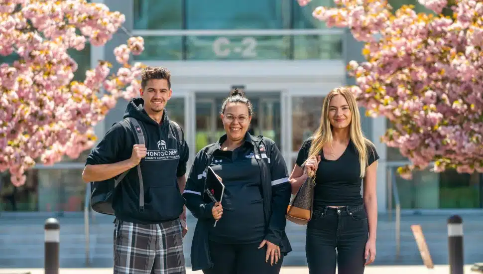 Three Montgomery County Community College students smiling and standing in front of a campus building, framed by blooming cherry blossom trees on a sunny day.