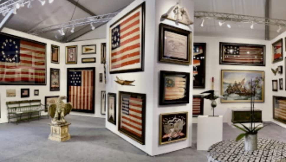 Exhibit booth at an antiques show displaying framed historic American flags, patriotic artwork, and Americana artifacts, including a carved eagle sculpture and vintage furnishings.