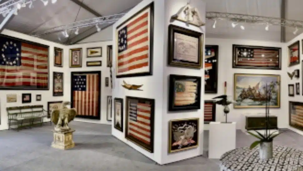 Exhibit booth at an antiques show displaying framed historic American flags, patriotic artwork, and Americana artifacts, including a carved eagle sculpture and vintage furnishings.