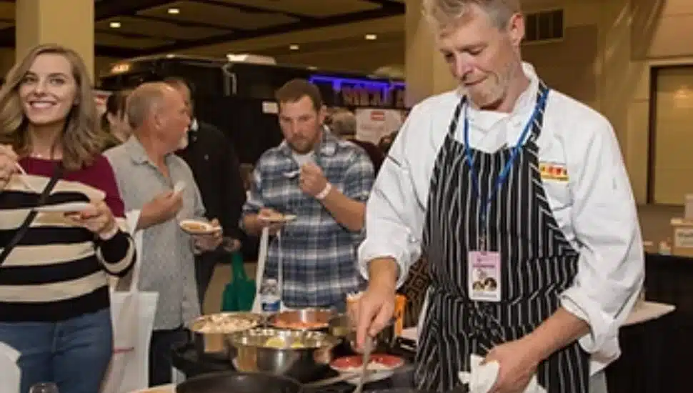 A chef in a striped apron prepares food at a tasting station while smiling attendees sample dishes at the TASTE! Philadelphia Festival. The scene is lively, with guests enjoying gourmet bites in a festive indoor setting.
