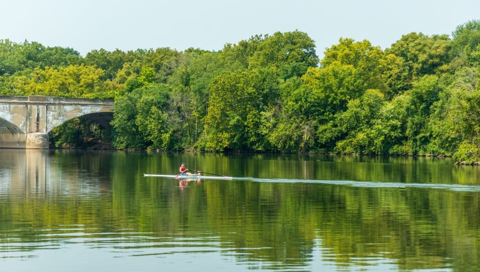 A man and a woman are rowing a boat in Schuylkill river. The water is calm and the trees in the background are lush and green. The scene is peaceful and serene.