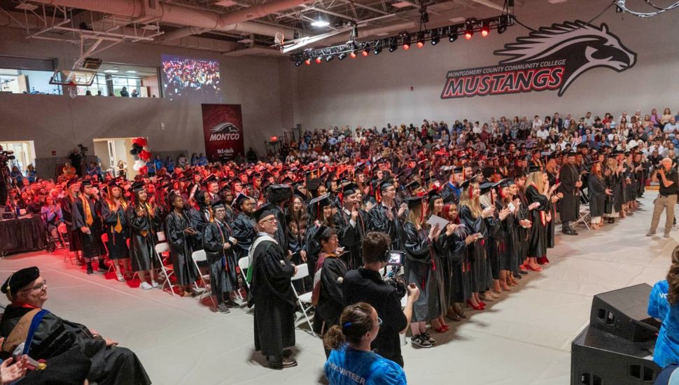 Montgomery County Community College graduation ceremony with graduates in black caps and gowns standing and applauding in a large gymnasium filled with faculty, families, and friends. The MCCC Mustangs logo is displayed on the wall, and red lighting adds a festive atmosphere.