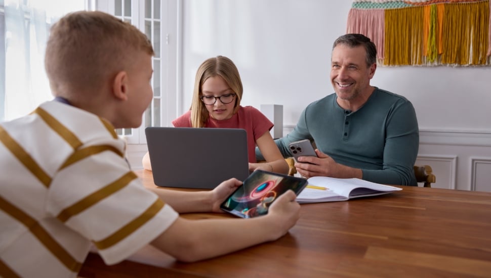 A family sitting at a table with a cellphone, laptop, IPad, and notebook and pencil.