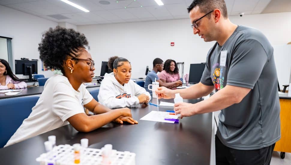 A male instructor demonstrates a science experiment to two attentive students at a lab table in a modern classroom. The students, one wearing glasses and the other in a white hoodie, observe as he uses a pipette over a worksheet. In the foreground, a test tube rack holds colorful liquids. Other students are seated in the background, working at computers and engaging in discussion. The setting is bright, organized, and designed for interactive STEM learning.