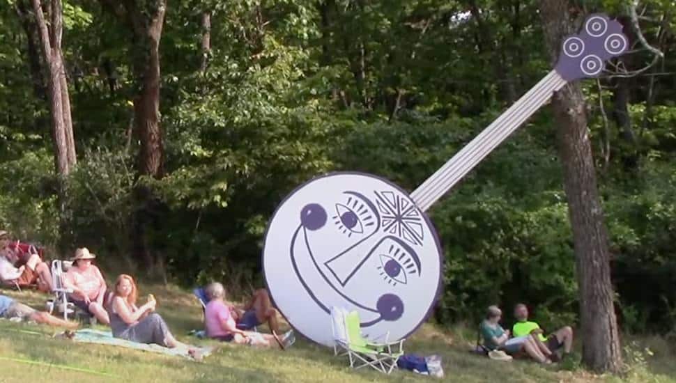 A large cutout of the Philadelphia Folk Festival Banjo leans against a tree with campers sitting around the bottom.