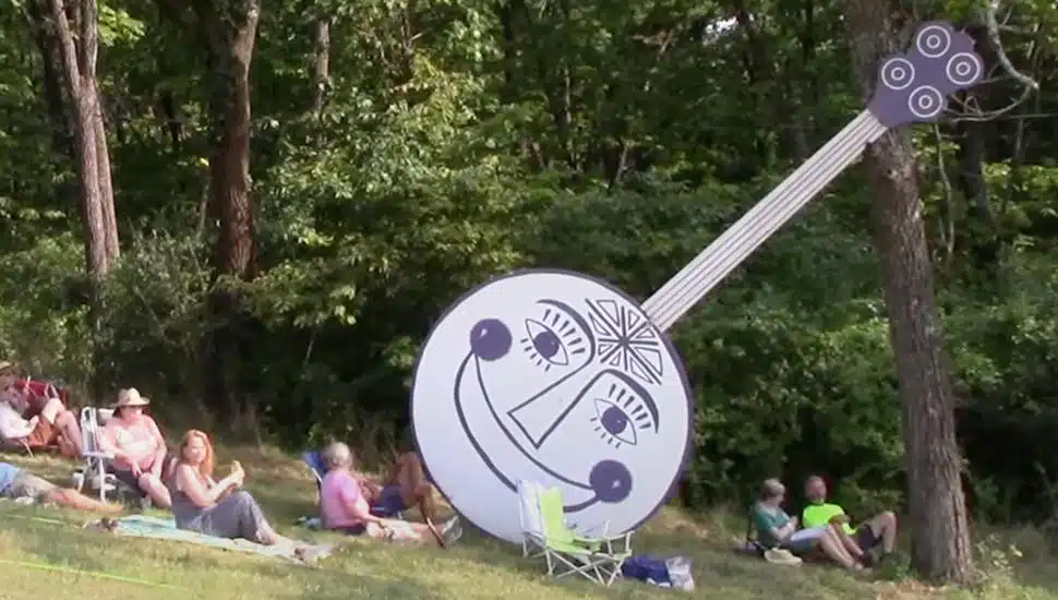 A large cutout of the Philadelphia Folk Festival Banjo leans against a tree with campers sitting around the bottom.