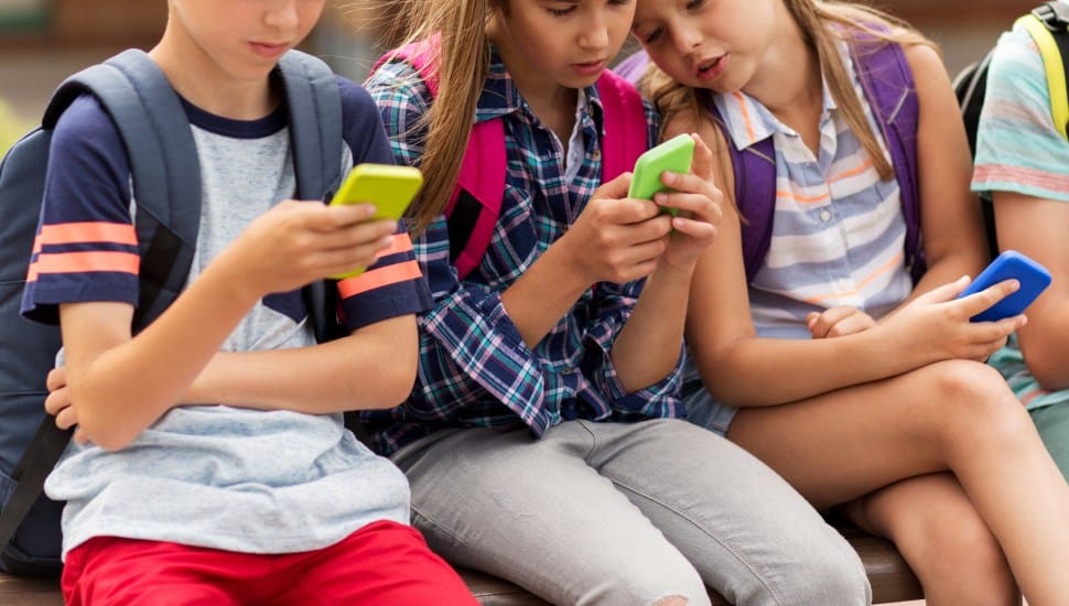 A group of elementary school students with smartphones and backpacks sitting on bench outdoors.