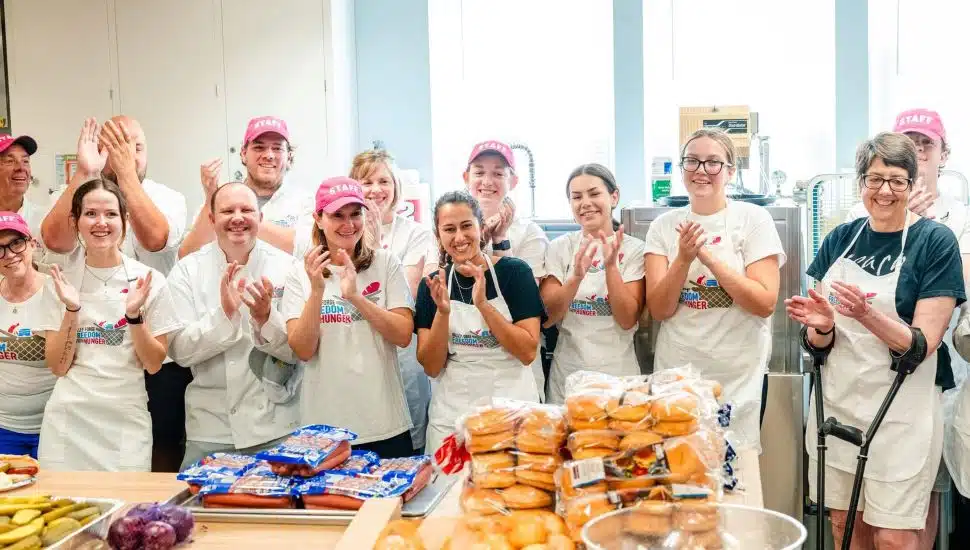 Group of Valley Forge Tourism & Convention Board volunteers in aprons and staff hats smiling and clapping in a kitchen during the Freedom from Hunger Drive kickoff, with food supplies like hot dogs and rolls on the counter.