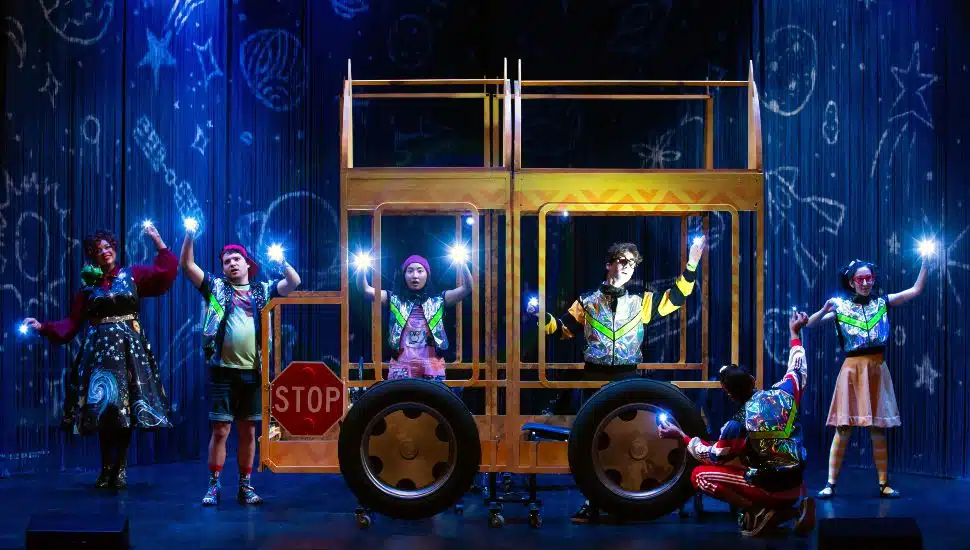 Cast of TheaterWorksUSA’s “The Magic School Bus: Lost in the Solar System” performing on stage, with actors in colorful costumes standing around a yellow school bus frame, holding glowing lights, against a backdrop decorated with space-themed drawings.