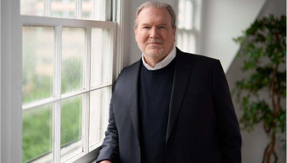 An older man in a dark suit jacket and sweater stands by a bright window, looking at the camera with a calm, confident expression.