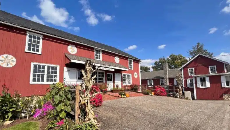 Exterior view of the Artisan Village at Bird-in-Hand, featuring a large red barn-style building with quilt block decorations, surrounded by colorful flowers, fall décor, and smaller red outbuildings under a bright blue sky.