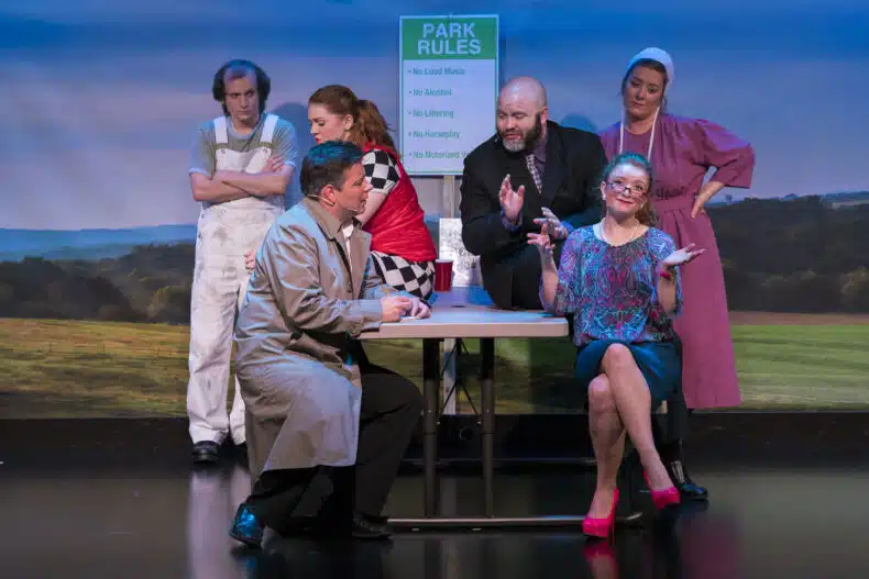 A group of six actors performing on stage in the play Stolen at Bird-in-Hand Stage. They are gathered around a picnic-style table under a sign that reads “Park Rules,” engaged in animated conversation and gestures, with colorful costumes representing a mix of modern and Amish influences.