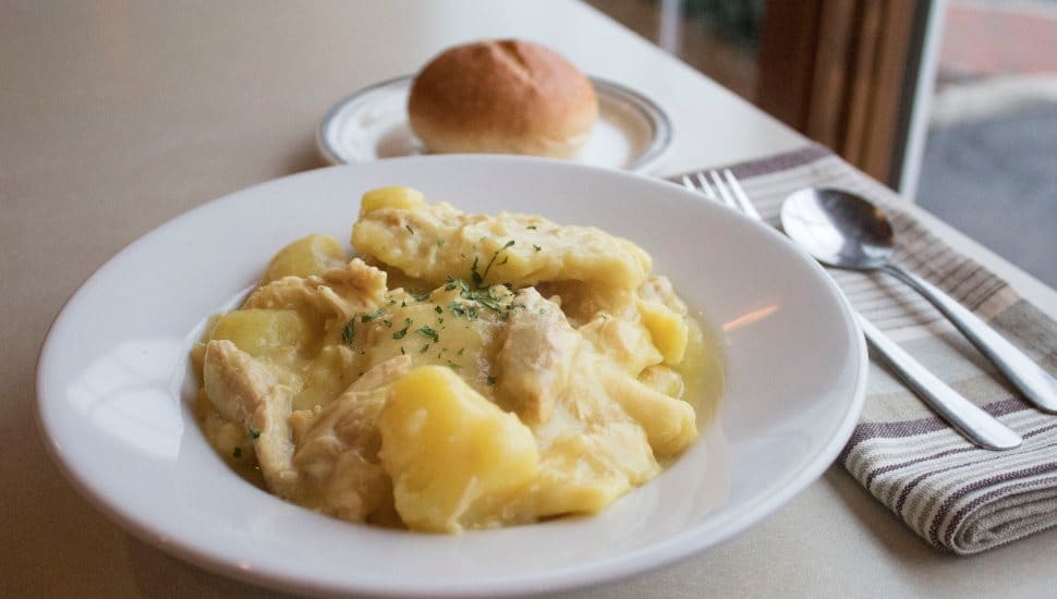 Close-up of a white bowl filled with Pennsylvania Dutch chicken pot pie featuring chunks of chicken, potatoes, and wide noodles in a savory broth, served with a dinner roll on the side.