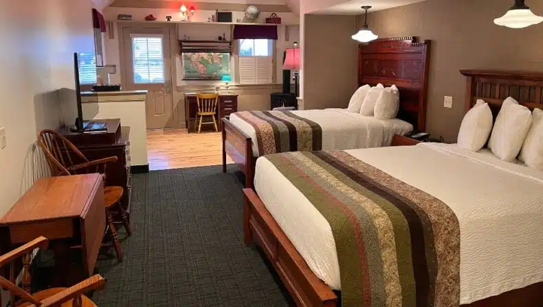 Hotel room at Bird-in-Hand Family Inn Schoolhouse Suite with two queen beds, quilted runners, wooden headboards, a desk with a map on the wall, warm lighting, and classic Amish-country style furnishings.