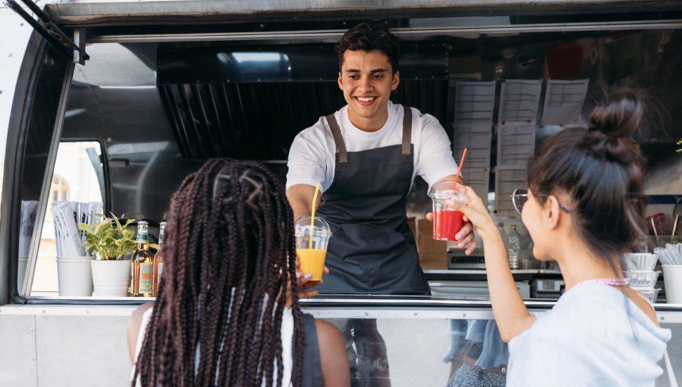 Back view of two females buying drinks from a male owner at a food truck.